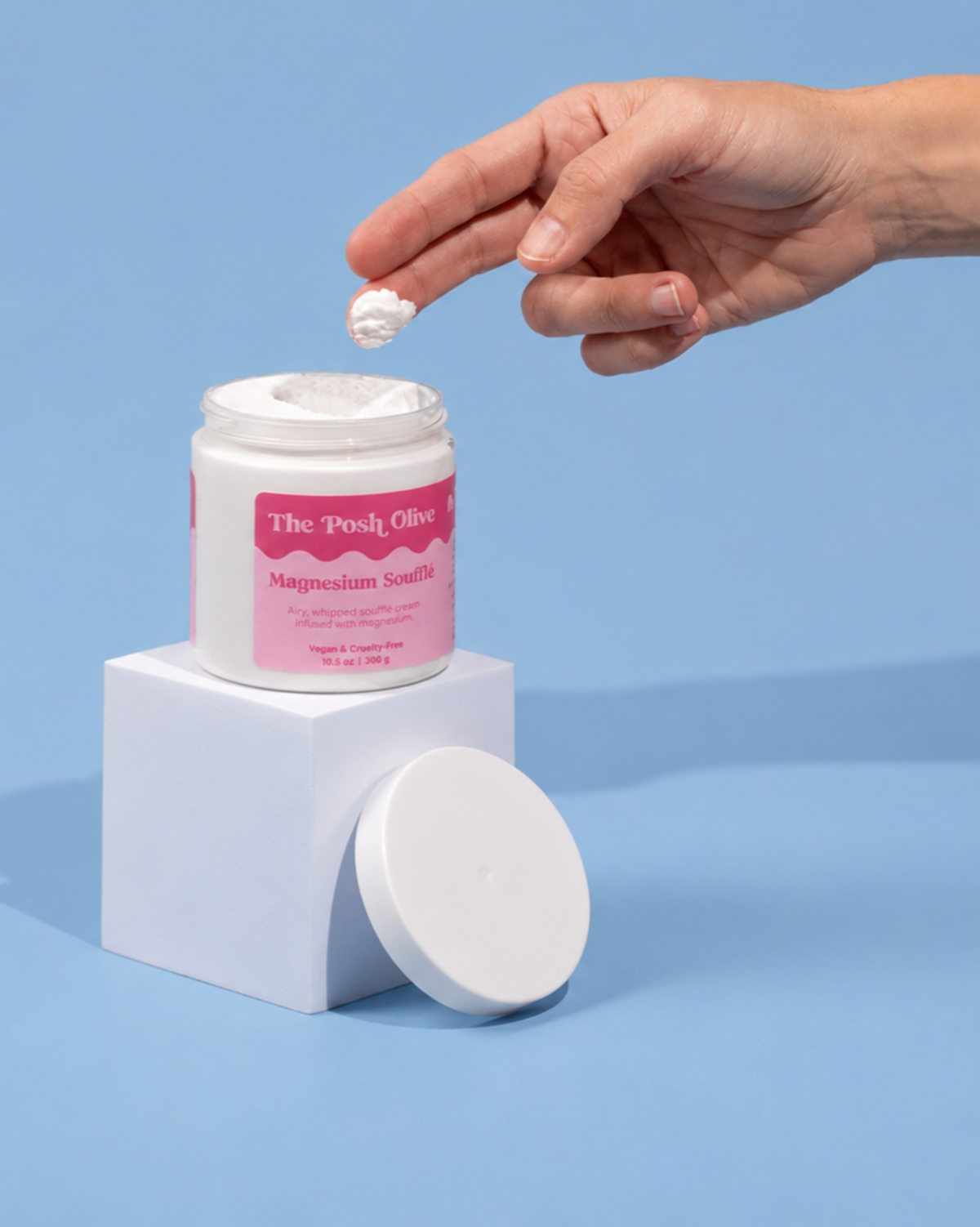 A hand holds a small dollop of white cream above an open white jar with a pink label reading 'The Posh Office Magnesium Scalp.' The jar sits on a white pedestal against a light blue background, with the lid resting beside it.