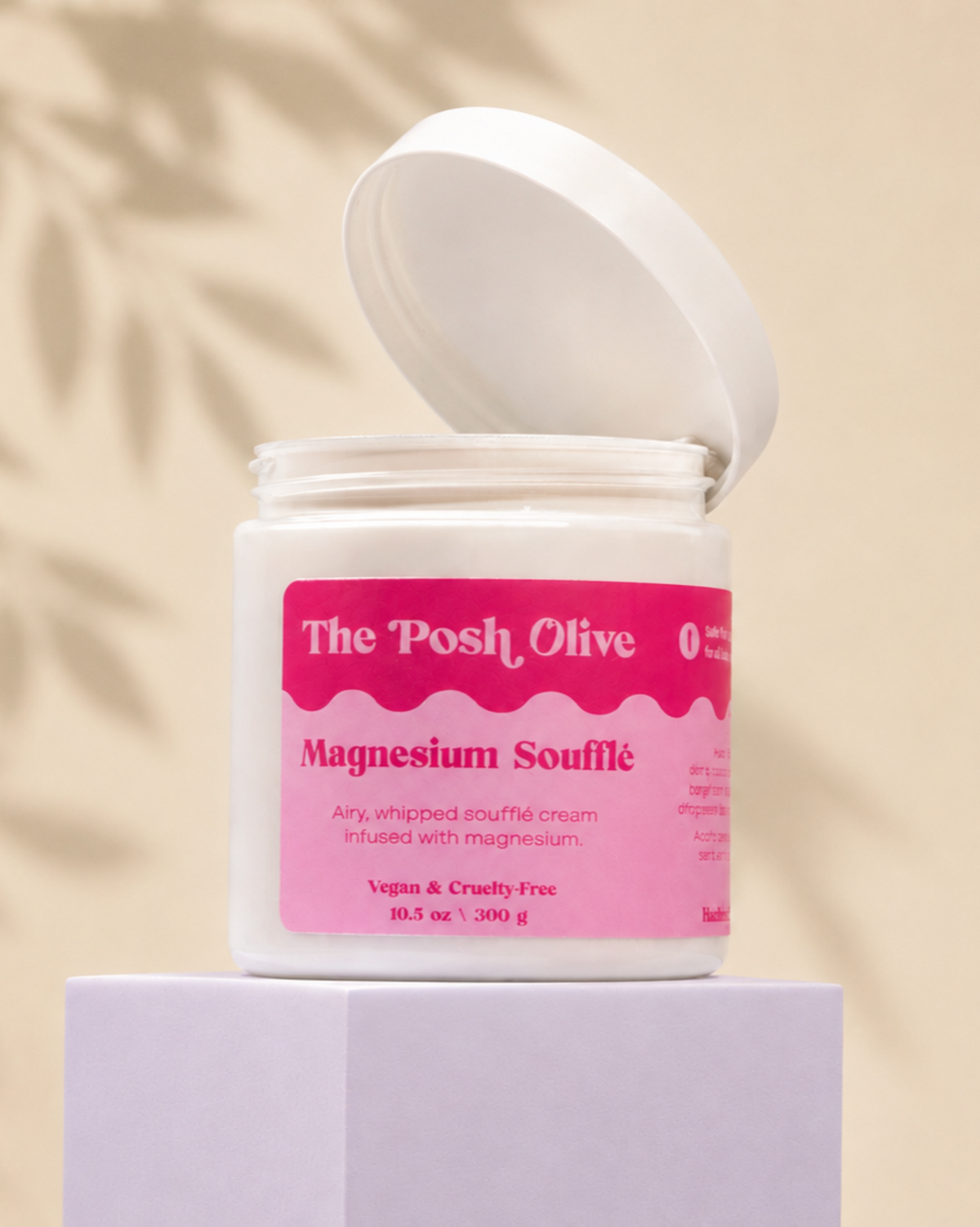 A white jar of The Posh Olive Magnesium Soufflé whipped cream, with its lid open and resting on a white pedestal against a warm beige background with soft leaf shadows.