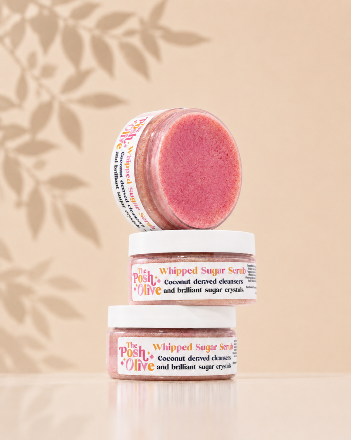 Three small jars of whipped sugar scrub stacked on a reflective surface against a warm beige background with soft leaf shadows. The top jar is tipped over to show the pink scrub inside, and the labels read 'The Posh Olive' and 'Whipped Sugar Scrub'.