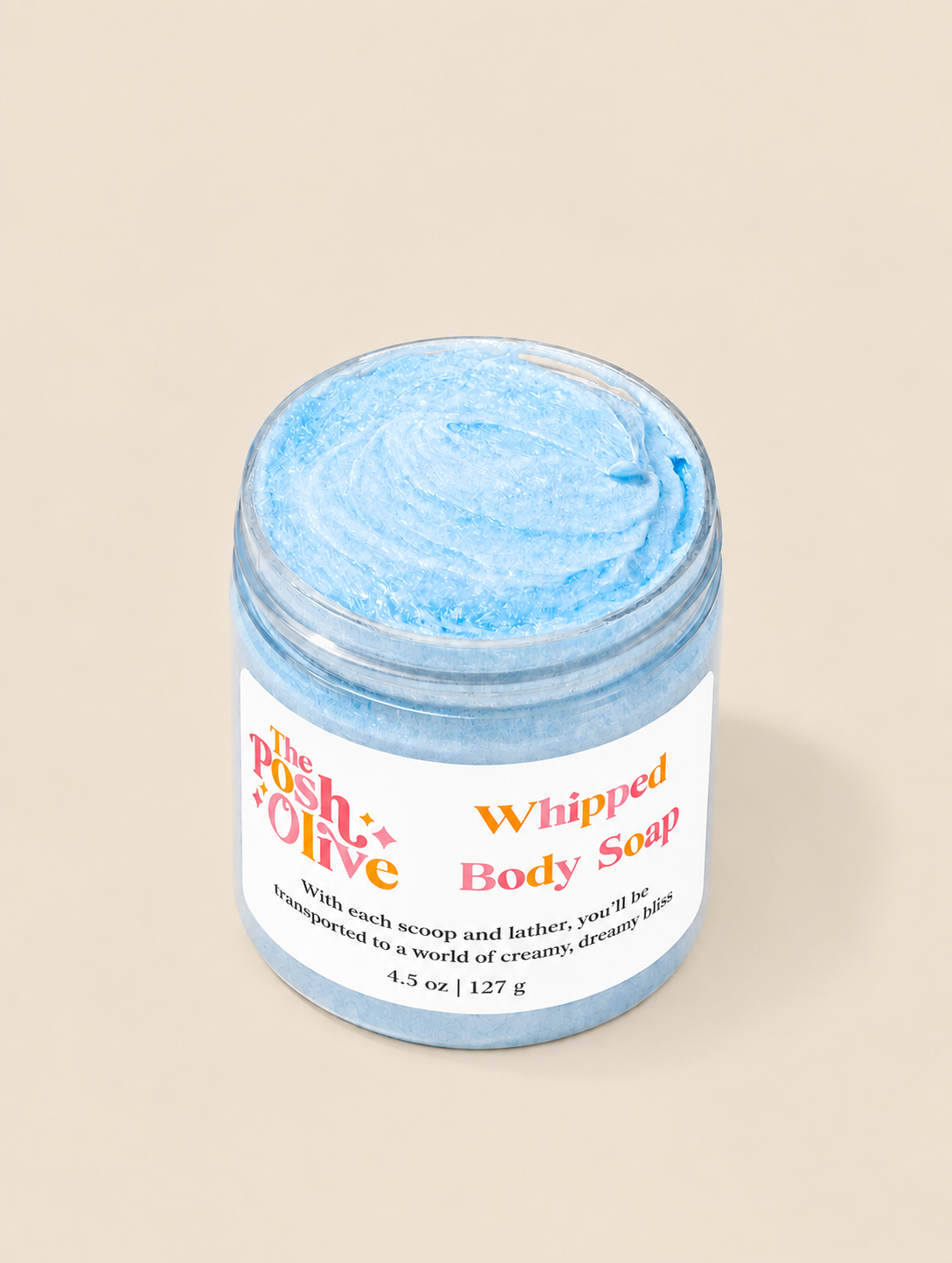 A jar of whipped body soap with a light blue whipped texture, featuring a white label with orange and red text, set against a solid beige background.