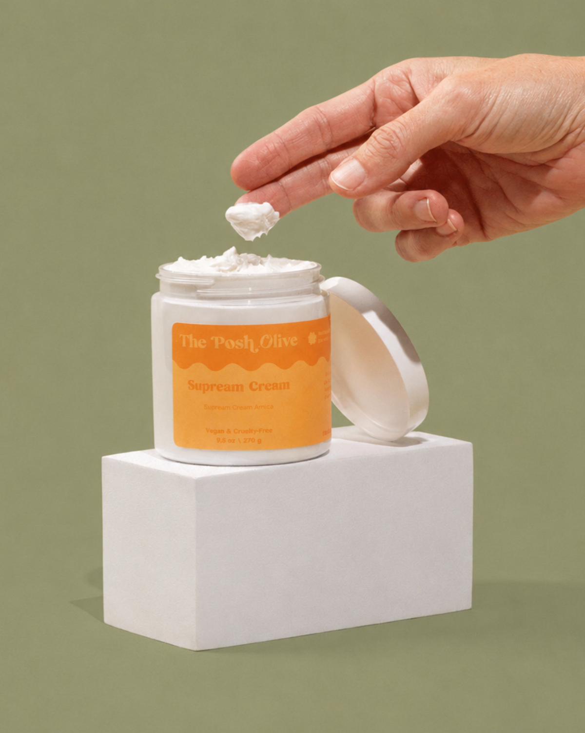 A hand scoops white cream from an open jar labeled 'The Pastel Olive Supream Cream' set on a white pedestal against a sage green background.