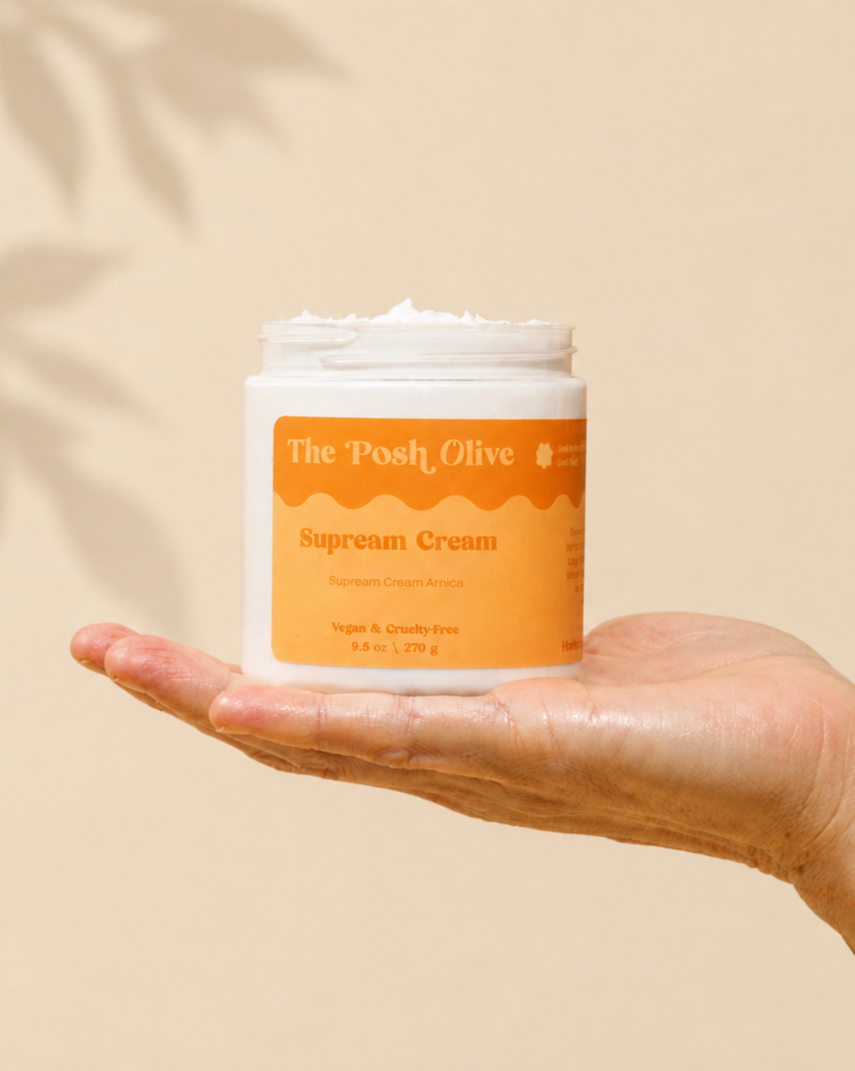 A hand holds a white jar of The Posh Olive Supream Cream against a warm beige background with soft leaf shadows.